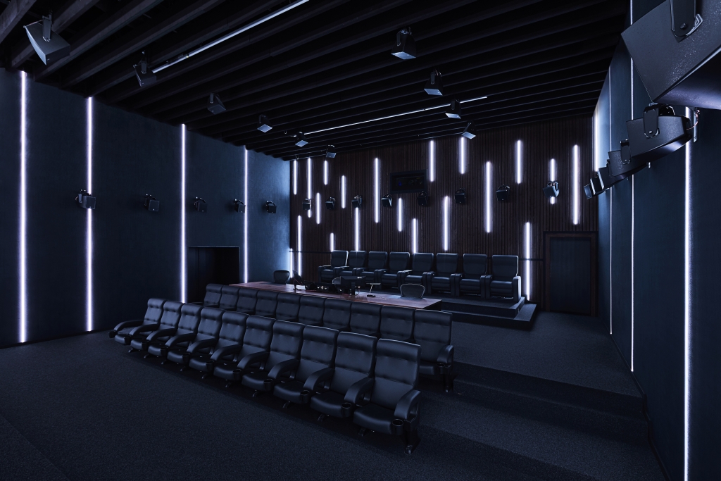 Ein modernes, dunkel beleuchtetes Auditorium mit zwei Reihen schlichter schwarzer Sitze, die auf eine Bühne ausgerichtet sind. Vertikale LED-Lichtleisten schmücken die Wände, und an der Decke und den Wänden sind Lautsprecher angebracht, die eine High-Tech-Atmosphäre schaffen.