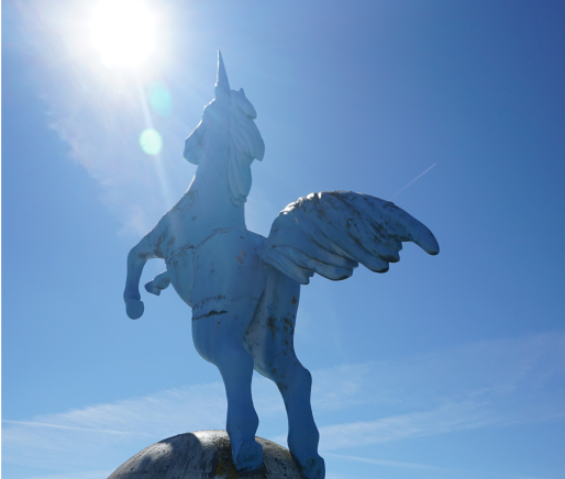 Eine blaue Statue eines geflügelten Einhorns steht auf einer Kugel und blickt in die helle Sonne unter einem klaren blauen Himmel. Das Sonnenlicht erzeugt ein starkes Streulicht, das die Statue teilweise in den Schatten wirft.