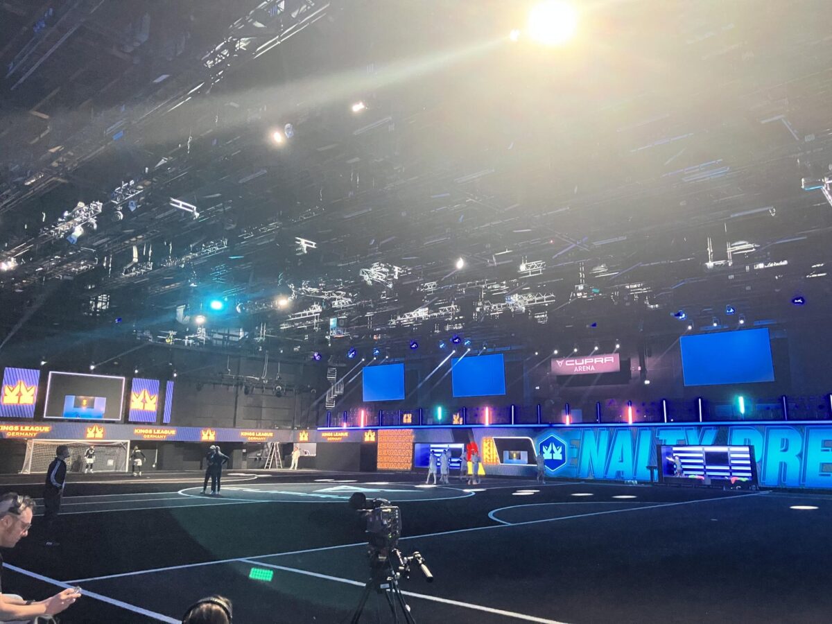 Eine große Sporthalle ist mit blauen und violetten Lichtern beleuchtet. Nur wenige Menschen stehen auf dem schwarzen Spielfeld, und es sind Kameras aufgestellt. Digitale Bildschirme und Banner mit Kronensymbolen und Texten schmücken die Wände.