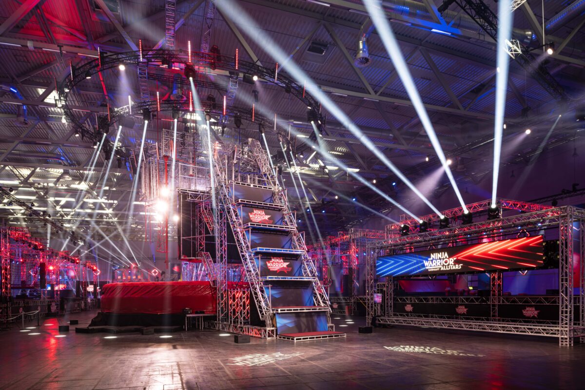 Ein hell beleuchteter Indoor-Hindernisparcours von Ninja Warrior mit hohen Metallstrukturen, Rampen, Plattformen und bunten Lichtern in Rot, Blau und Weiß, mit Scheinwerfern und dem Ninja Warrior-Logo.