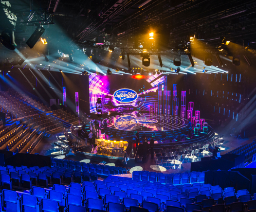 Ein hell erleuchtetes Fernsehstudio mit einer großen, kreisförmigen Bühne mit bunten Lichtern und dem "SuperStar"-Logo auf einem großen Bildschirm. Reihen von leeren Zuschauersitzen umgeben die Bühne, und verschiedene Bühnengeräte sind zu sehen.