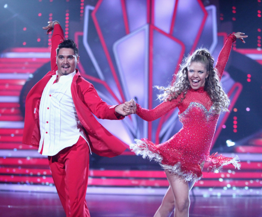 Ein Mann und eine Frau in passenden knallroten Kostümen führen auf einer hell erleuchteten Bühne eine energiegeladene Tanznummer auf, halten sich an den Händen und lächeln mit jeweils einem erhobenen Arm.