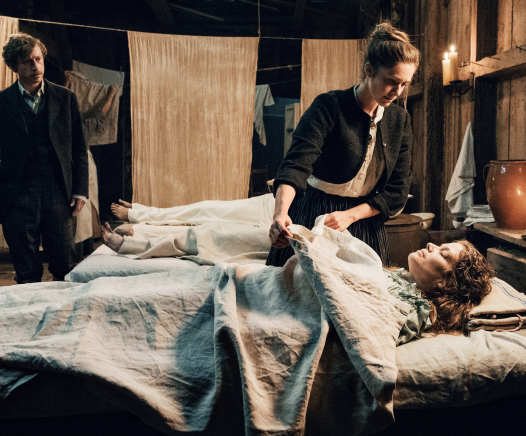 Eine Frau kümmert sich um eine andere Frau, die im Bett liegt, und deckt sie mit einer Decke zu, während ein Mann in der Nähe in einem schwach beleuchteten, rustikalen Raum mit Kerzen und aufgehängter Wäsche steht.