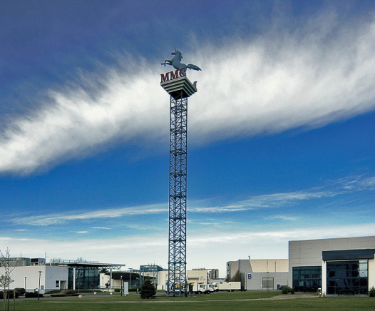 Ein hoher Metallturm mit einer fliegenden Pferdestatue und einem MMC-Zeichen an der Spitze steht vor modernen Industriegebäuden unter einem blauen Himmel mit Wolkenfetzen.