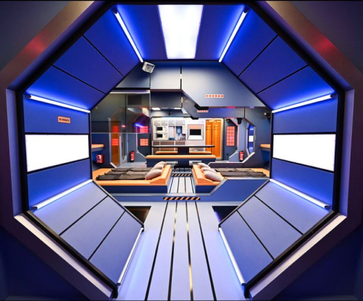 Ein futuristisches, sechseckiges Zimmer mit blauen Wänden, Neonbeleuchtung, zwei Betten und einer modernen Küche im Hintergrund, die an das Innere eines Science-Fiction-Raumschiffs erinnert.