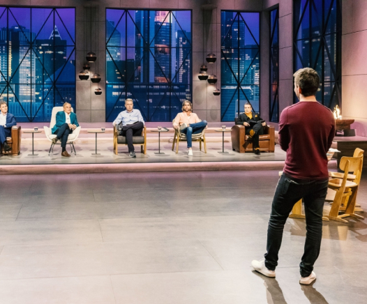 In einem hell erleuchteten Fernsehstudio mit großen Fenstern, die eine nächtliche Stadtlandschaft zeigen, steht eine Person und präsentiert fünf sitzenden Juroren.