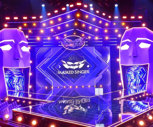 Die hell erleuchtete Bühne für The Masked Singer ist mit großen geometrischen Gesichtsskulpturen auf beiden Seiten, lebhafter blauer und violetter Beleuchtung und dem Logo der Show in der Mitte ausgestattet.