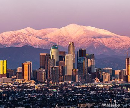 Skyline der Innenstadt von Los Angeles bei Sonnenuntergang mit hohen Wolkenkratzern im Vordergrund und schneebedeckten Bergen im Hintergrund unter einem rosafarbenen Himmel.