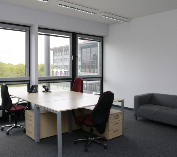 Ein modernes Büro mit drei Stühlen um einen großen Schreibtisch, einem Telefon auf dem Schreibtisch, großen Fenstern mit Blick auf ein anderes Gebäude und Bäume und einem grauen Sofa vor einer weißen Wand.
