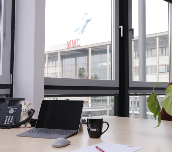 Ein moderner Bürotisch mit Laptop, Telefon, Kaffeetasse, Pflanze und Notizblock. Große Fenster zeigen ein Gebäude mit MMC-Beschriftung und einer weißen Pferdestatue auf dem Dach.
