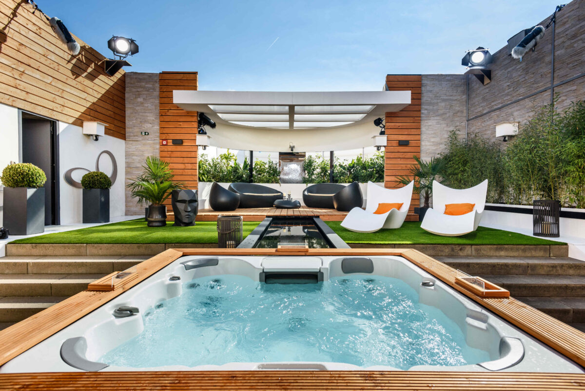 Auf der modernen Dachterrasse gibt es einen Whirlpool, weiße Liegestühle mit orangefarbenen Kissen, schwarze Sitzgelegenheiten, grünen Kunstrasen, Topfpflanzen und Holzverkleidung unter freiem Himmel.