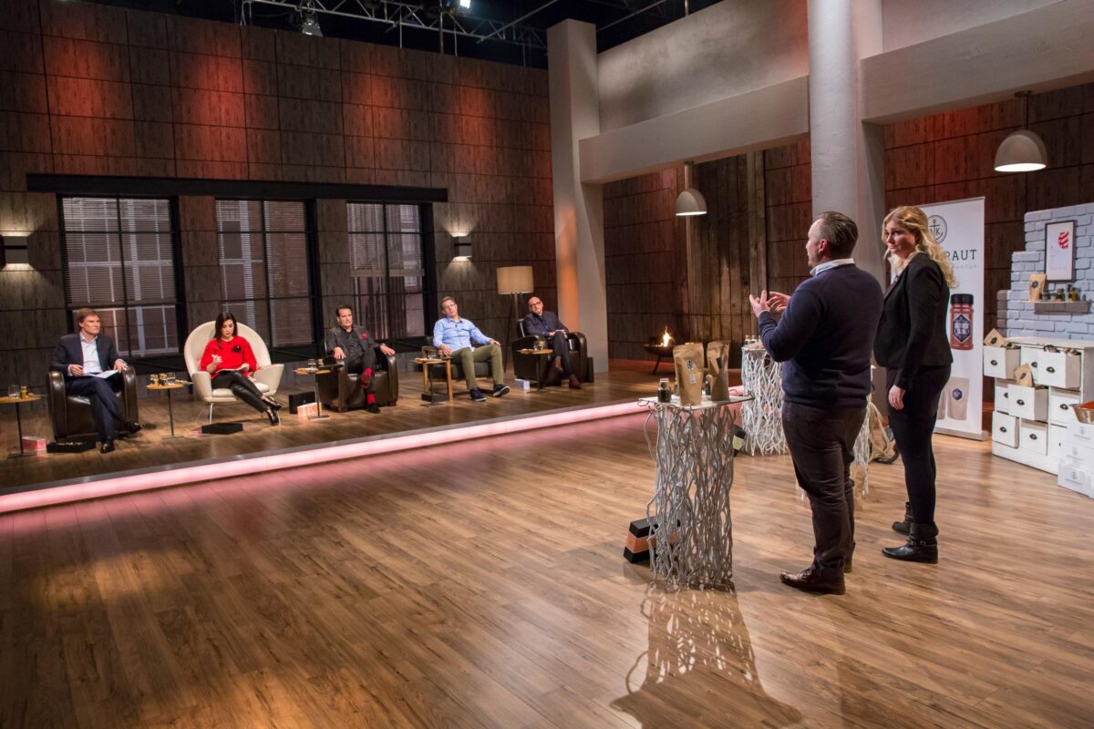 Zwei Personen präsentieren ein Produkt vor fünf sitzenden Jurymitgliedern in einem hell erleuchteten Fernsehstudio, ähnlich wie bei einer Business-Pitch-Show. Der Raum ist mit Holzböden, modernem Dekor und ausgestellten Produktmustern ausgestattet.