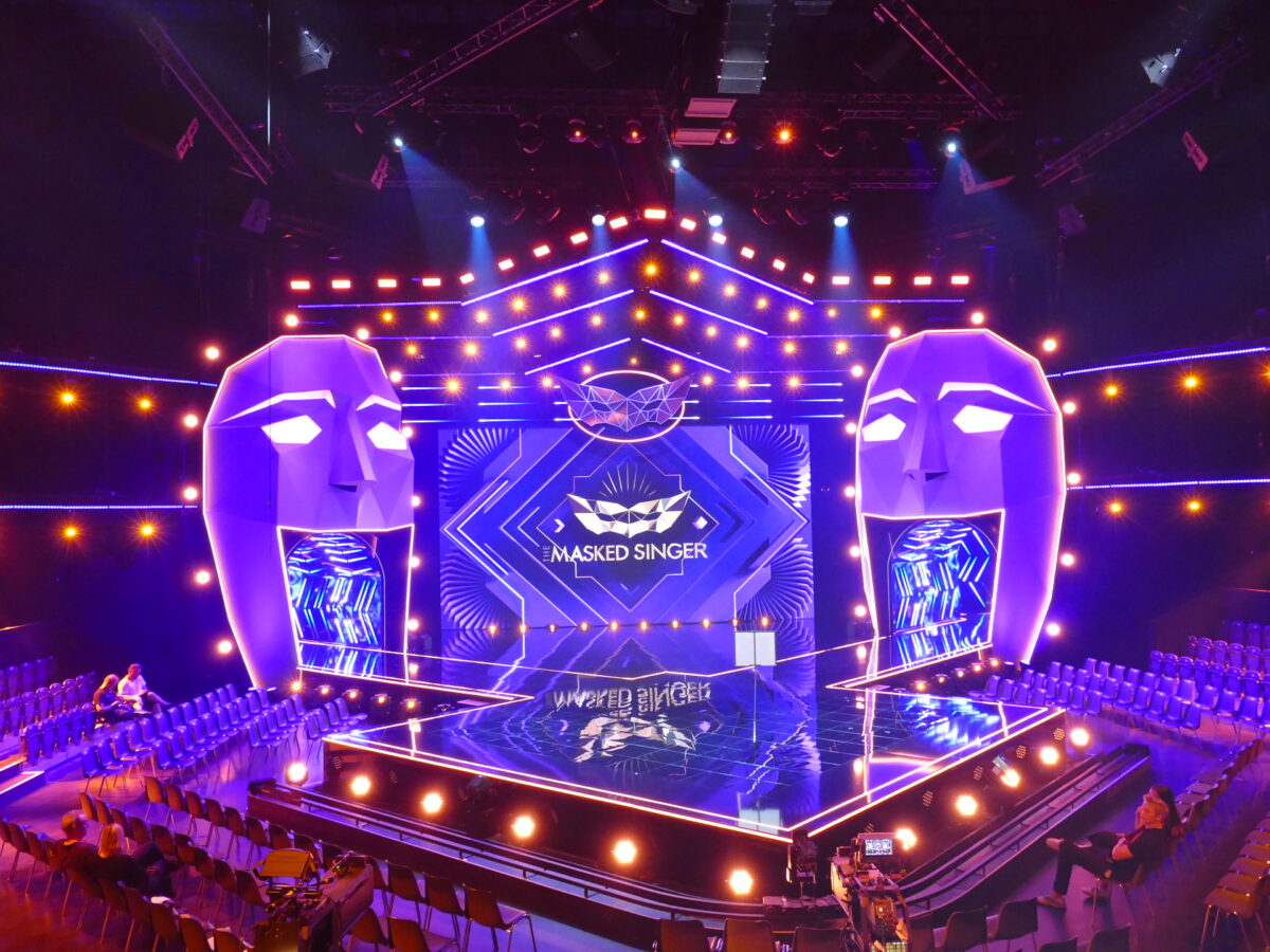 Auf der hell erleuchteten Bühne der Masked Singer stehen zwei große Maskenskulpturen, leuchtend lila und orangefarbene Beleuchtung, geometrische Kulissen und Reihen leerer Zuschauerplätze, die eine reflektierende Hauptbühne umgeben.