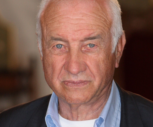 Ein älterer Mann mit kurzen weißen Haaren und blauen Augen blickt in die Kamera. Er trägt ein hellblaues Hemd mit Kragen und eine dunkle Jacke und hat einen neutralen Gesichtsausdruck. Der Hintergrund ist sanft verschwommen.