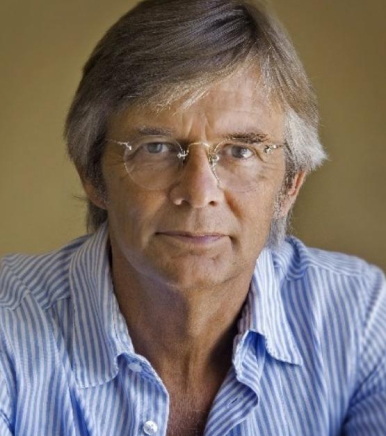 Ein Mann mittleren Alters mit grauem Haar und Brille trägt ein blau-weiß gestreiftes Button-up-Hemd. Er blickt direkt in die Kamera vor einem schlichten, neutralen Hintergrund.