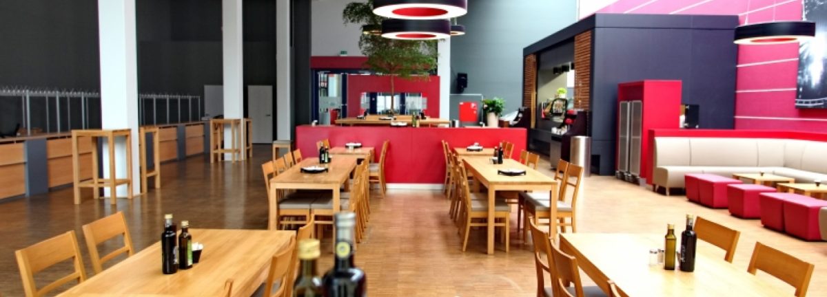 Ein modernes, geräumiges Restaurant mit Holztischen und -stühlen, roten Akzenten, Flaschen auf Tischen, einem Barbereich und einer Mischung aus Steh- und Thekenplätzen. Helle Beleuchtung und große Fenster schaffen eine einladende Atmosphäre.