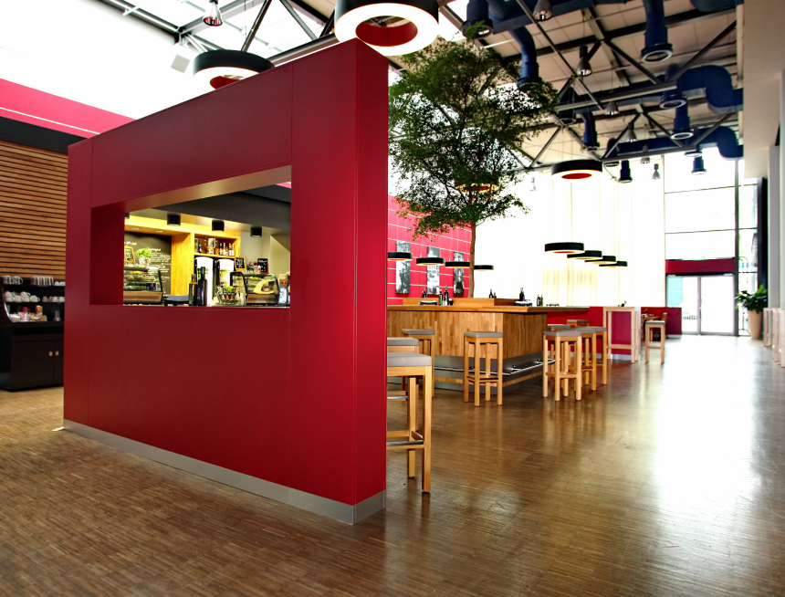 Modernes Café-Interieur mit roten Wänden, Holztischen und -hockern, großen Fenstern, die natürliches Licht hereinlassen, und einem Baum in der Mitte. Der Raum hat eine geräumige, luftige Atmosphäre mit industriellen Deckendetails.