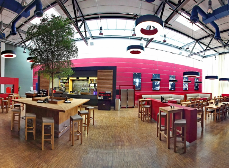 Moderne Cafeteria mit Holztischen und -hockern, einem großen Topfbaum als Mittelstück, roten Akzentwänden und Deckenleuchten. Der Raum ist offen und geräumig gestaltet, mit einer Theke und Regalen im Hintergrund.
