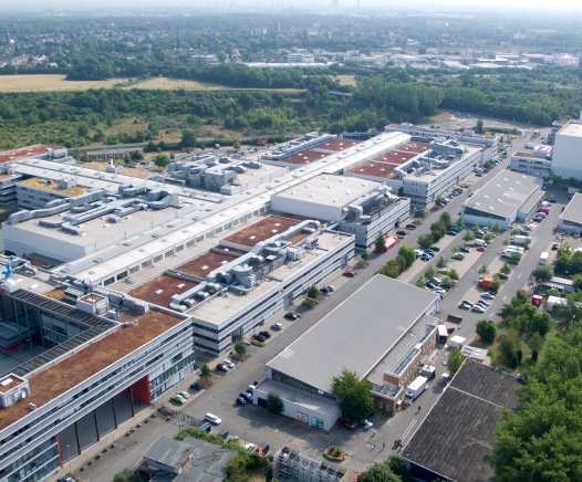 Luftaufnahme eines großen Industriekomplexes mit mehreren Flachdachgebäuden, Parkplätzen und Straßen, umgeben von Bäumen und Grünflächen, mit einer dunstigen Stadtlandschaft in der Ferne.
