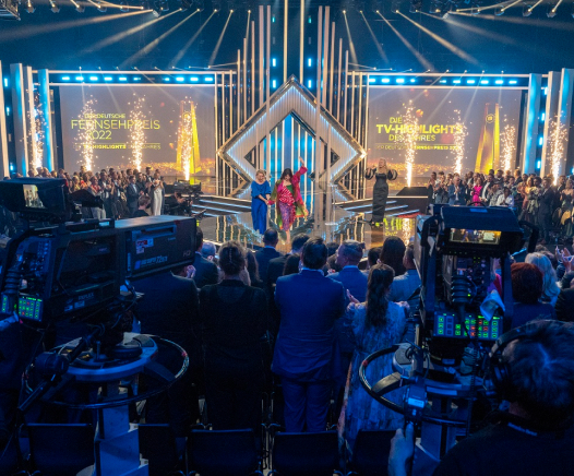 Das Publikum applaudiert bei einer hell erleuchteten Preisverleihung, bei der die Gastgeber oder die Gewinner auf der Bühne stehen. Auf großen Bildschirmen werden die TV-Highlights und der Fernsehpreis 2022 gezeigt. Im Vordergrund sind Kameras und Filmmaterial zu sehen.