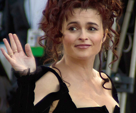 Eine Frau mit lockigem, kastanienbraunem Haar in einer Hochsteckfrisur und einem schwarzen Kleid lächelt und winkt mit ihrer linken Hand bei einer Veranstaltung im Freien.