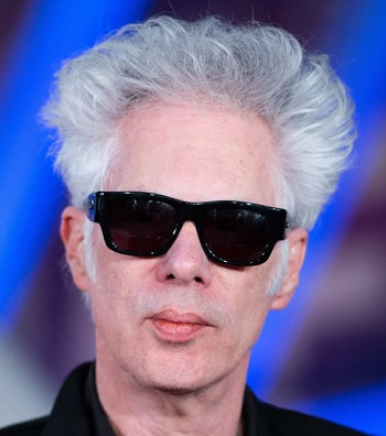 Eine Person mit heller Haut, weißem, voluminösem Haar und einer schwarzen Sonnenbrille blickt direkt in die Kamera. Der Hintergrund ist blau und lila und wirkt unscharf.