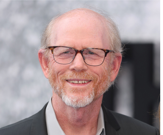 Ein älterer Mann mit heller Haut, kahlem Kopf, grauem und rötlichem Bart und Brille lächelt, während er ein graues Hemd und einen dunklen Blazer trägt und vor einem verschwommenen Hintergrund steht.