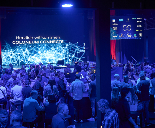 Eine große Gruppe von Menschen versammelt sich in einem schwach beleuchteten Veranstaltungsraum mit blauer Beleuchtung. Auf der Bühne zeigt ein Bildschirm "Herzlich willkommen COLONEUM CONNECTS" mit einer digitalen Netzwerkgrafik.