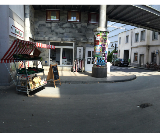 Ein kleiner Marktstand mit einer rot-weiß gestreiften Markise befindet sich neben einem Ladeneingang. In der Nähe steht eine mit bunten Plakaten bedeckte Säule unter einem Teildach, mit Gebäuden und einem geparkten Auto im Hintergrund.